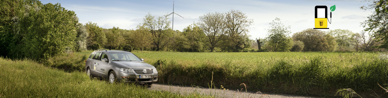 Ein PKW steht auf einer Wiese, im Hintergrund ist ein Windrad und Wald zu sehen.